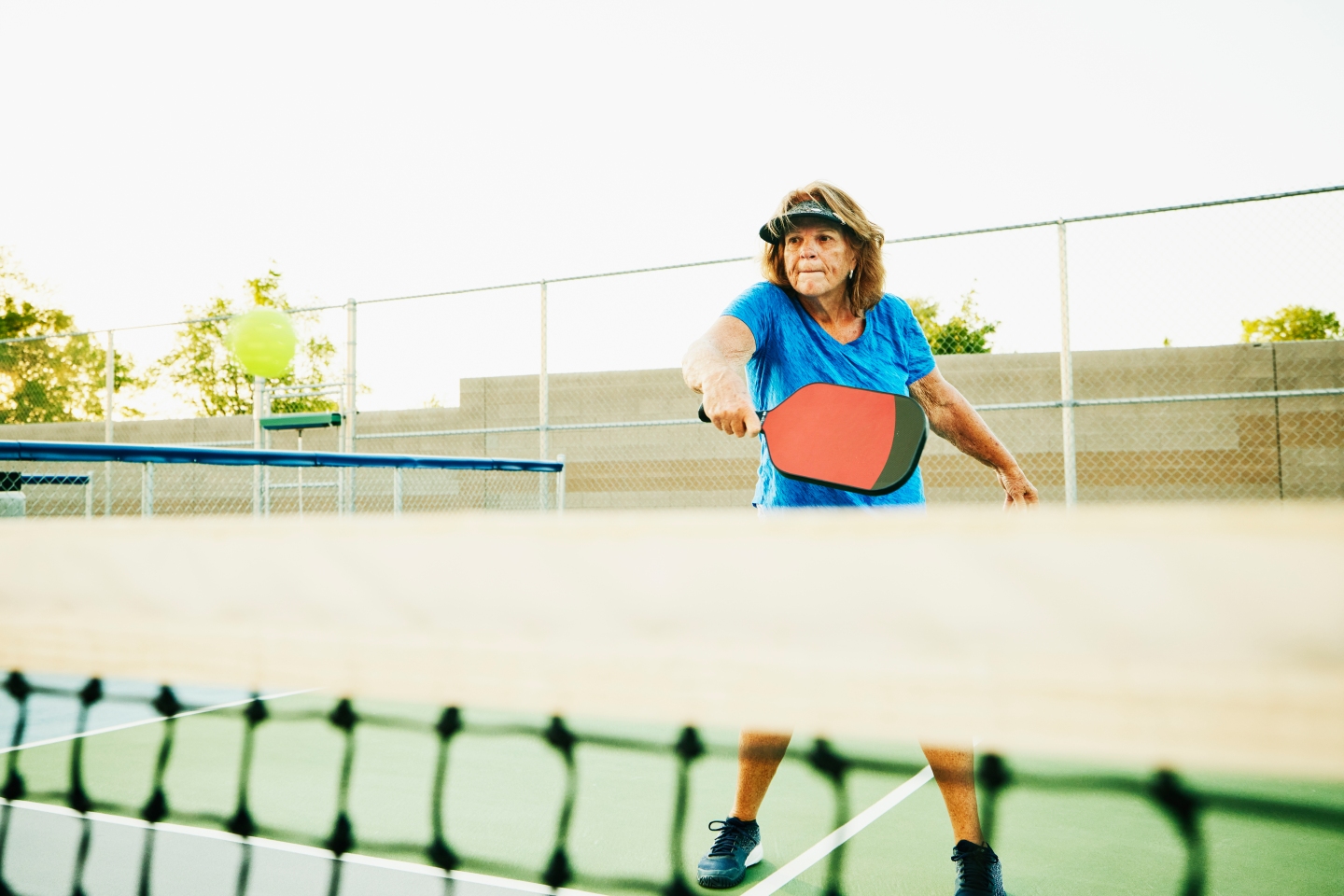 Wide shot of senior woman hitting backhand at net while playing pickleball on summer evening.